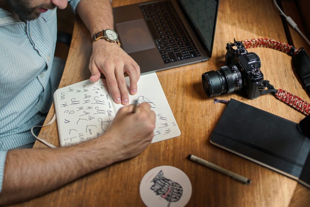 Image of a creative designer working in his sketchbook on a wooden desk with his camera next to him 